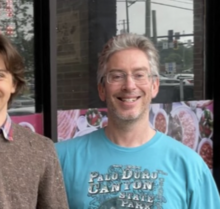 blurry image of Bryan Caplan wearing a light blue print t-shirt, grinning directly at camera