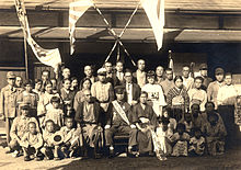 A family gathers around a young boy in a military uniform, surrounded by banners and flags. Some of the children also hold flags.