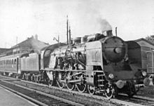 Photographie en noir et blanc d'un train à vapeur marquant l'arrêt en gare, locomotive légèrement fumante.