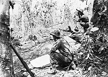 Black and white photo of a man wearing military uniform armed with a large gun lying down and aiming the weapon into dense bushland. Two other men in military uniform are crouched on either side of the prone man.