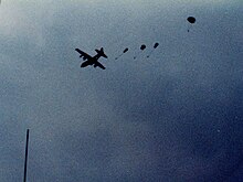 Four small parachutes can be seen coming out the back of a distant, silhouetted plane in flight. It is daytime, however the sky is dark and cloudy.