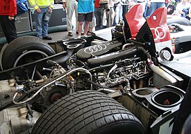 The Audi R10's exposed engine bay, viewed from the rear on a three-quarters angle. It shows the carbon-fibre intake plenums, which are emblazoned with the four-ring Audi logo and the letters "V12 TDI". Radiators are situated to the left and right of the engine, with air intakes for the rear wheels directly rearwards of them. The transmission and engine are both mounted longitudinally, with the former right behind the engine. The diesel particulate filters are immediately to the behind of the transmission.