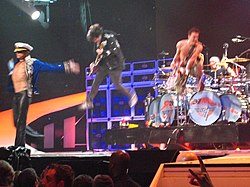 Van Halen performing onstage: David Lee Roth is wearing a sea captain's outfit with his arms outstretched, Woflgang Van Halen is jumping in the air with his bass guitar, Eddie Van Halen is also jumping while playing guitar, and Alex Van Halen is behind them drumming