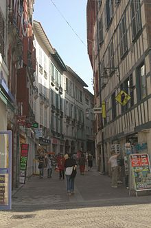 Photographie d’une rue étroite, s'incurvant vers la droite et bordée d’immeubles de 3 étages, aux façades blanches à pans de bois peints.