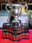 Silver bowl trophy with two large handles, mounted on a wide black plinth engraved with team names on silver plates.