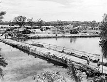 Black and white photo looking across a narrow body of water towards a town in which all of its buildings have been reduced to rubble.