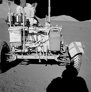 Cette vue de l'arrière du rover montre sur la droite la réparation effectuée sur le garde-boue. Le gravimètre est la boite grise à côté du garde-boue. Gene Cernan est assis à gauche. Immédiatement derrière lui se trouve le boitier contenant les charges explosives. Sur l'arrière du véhicule on distingue le râteau (à gauche) et l'écope et devant, un sac (SCB) dans lequel sont rangés les échantillons.