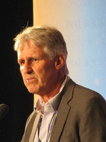 An older white man, with short white hair, in a beige jacket, stood in front of a presentation screen