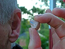 Close-up photograph of a person holding a hearing aid close to their left ear