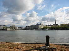 Vue d’une rivière, avec un quai en premier plan, des bâtiments en arrière plan et un ciel bleu envahi de cumulus.