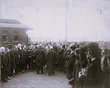 A black-and-white photograph of a crowd of men are standing in a semi-circle around Grover Cleveland. A train car is visible in the top-left corner of the photograph.