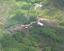 An overhead view of a set of buildings located in a dense jungle. A river runs to the left of the buildings, and moving trucks are seen to the right of the buildings. There is a dirt road leading from the buildings to other buildings.