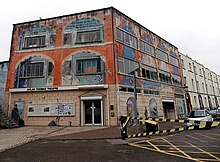 A wide three storied building with windows to the upper two stories and an entrance on the ground floor. A statue of Thomas sits outside.