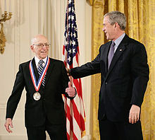 Un homme souriant en costume noir porte une médaille autour du cou, et un homme souriant en costume noir le congratule, avec sa main sur son épaule.