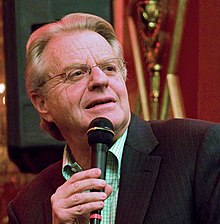 Headshot of a television host wearing a green shirt and a black suit jacket holding a microphone.