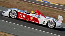 An Audi R10 in motion, viewed from its left side. The number 8 and the letters "TDI Power" are emblazoned on the side, next to the German flag and the Royal Dutch Shell logo. Frank Biela is at the wheel and is currently turning right.