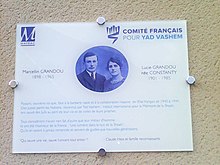 Plaque blanche portant en bleu la photo du couple Grandou jeunes, les logos de Mayrac et du Comité français Yad Vashem et une exhortation des passants à se souvenir.