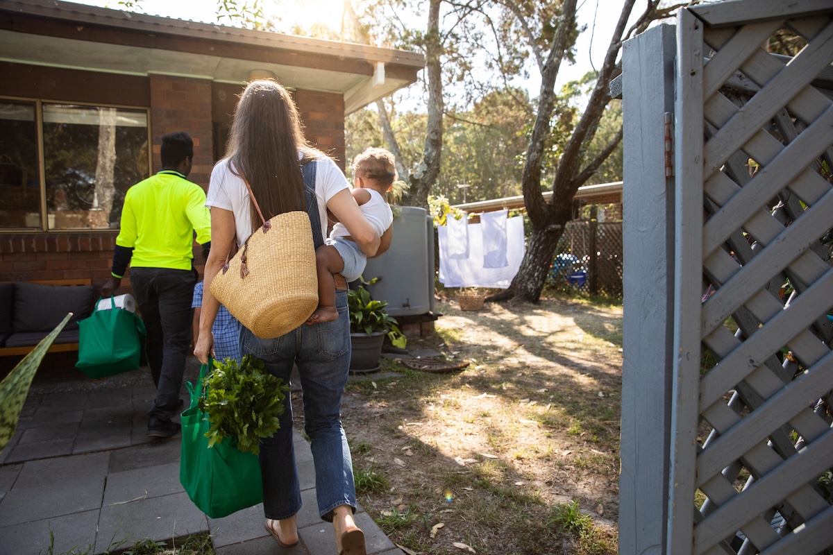 A family walking into their home with reusable bags, a rain barrel outside of the door, clothes hanging on a clothesline air drying in the backyard.