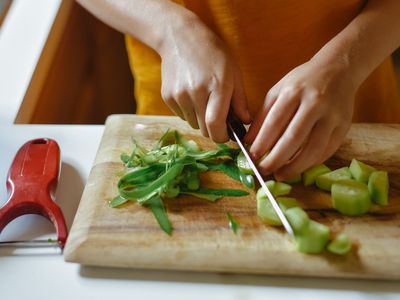 Person's hands cutting a cucumber with a knife. Peeler visible on the side of the cutting board.