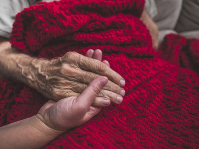 Closeup of a hand holding an elderly patient's hand over a red knit blanket.