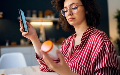 A woman holding up and reading a pill bottle in one hand with her smartphone in the other hand