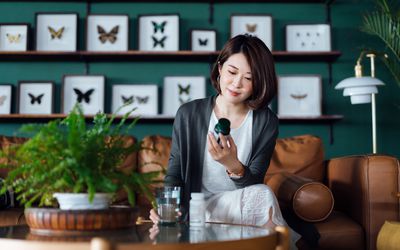 Young Asian woman taking medicines with a glass of water on the coffee table, reading a label of her medication at home.