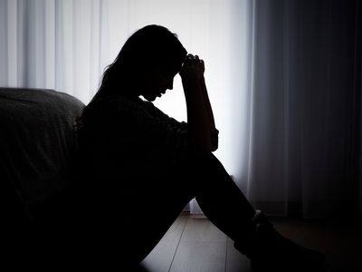 Woman with seasonal affective disorder sitting with her head in her hands on her bedroom floor, leaning against her bed