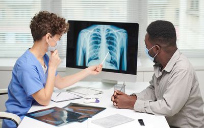 Side view shot of professional pulmonologist demonstrating x-ray shot to patient on computer screen and explaining something to him - stock photo