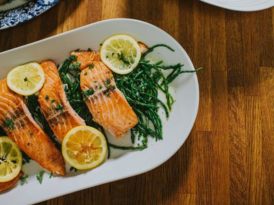 Top down image of four salmon darns on a white long oval serving ceramic plate on a wooden table. It rests on a bed of samphire. Lemon slices garnish the fish.