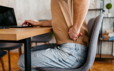 Man sitting in chair while working on a laptop with hand on back in pain