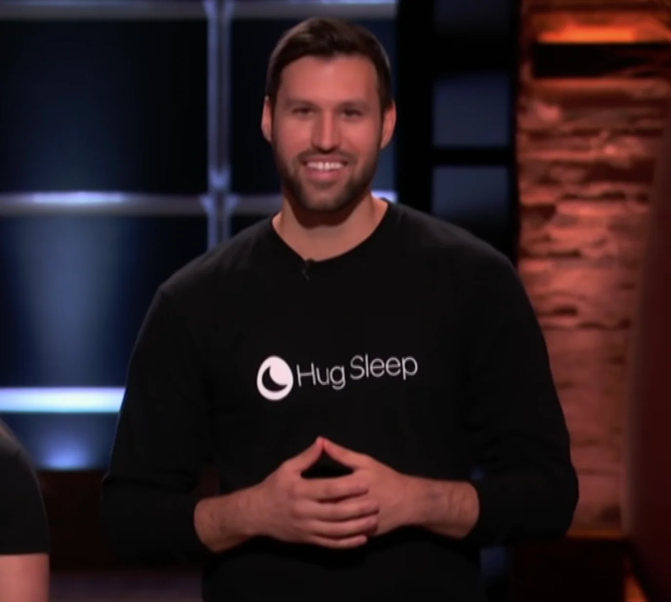 Smiling man in a black Hug Sleep shirt, standing on a stage in front of a dark background.