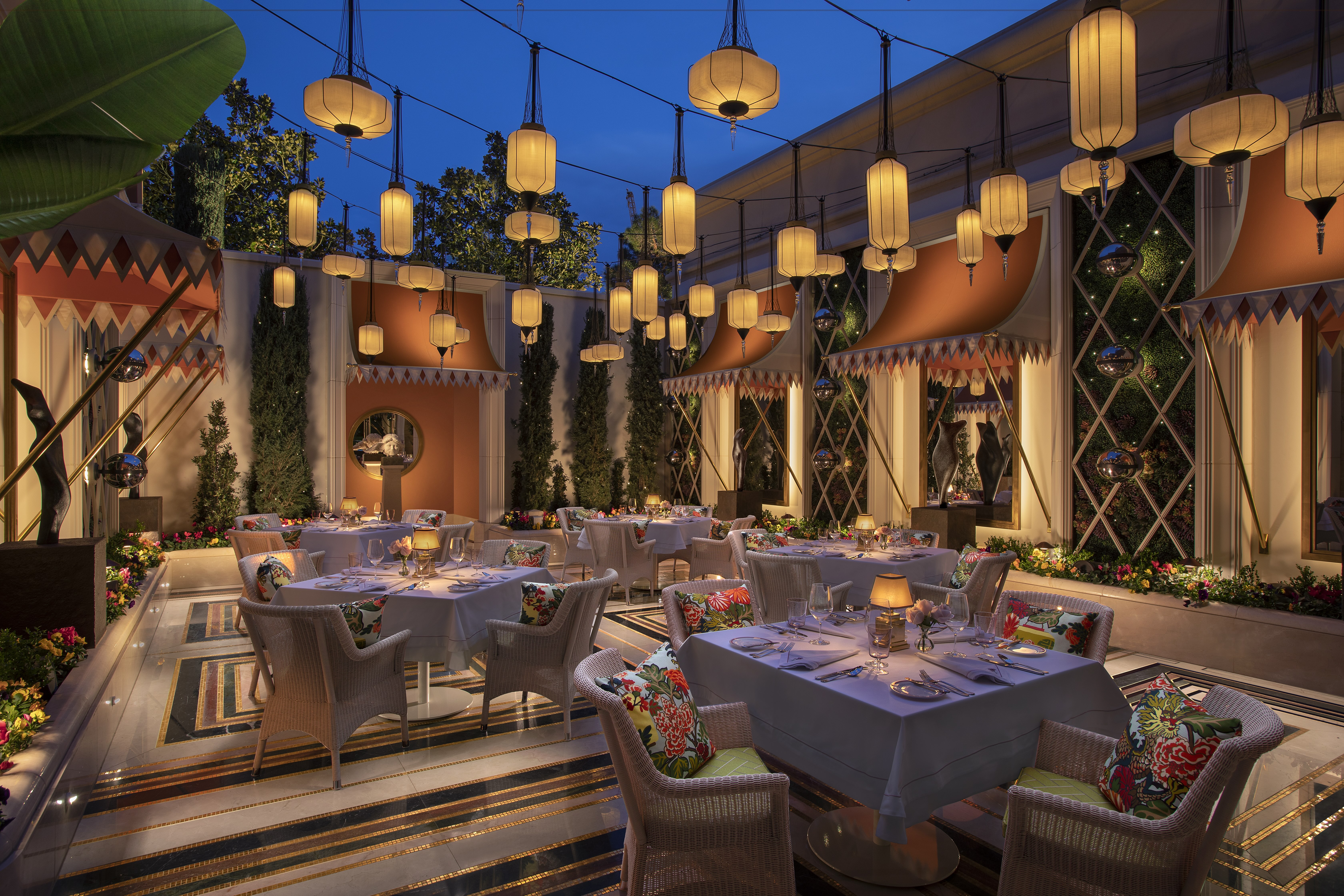 Strings on lanterns in different shapes hang above an outdoor patio with tables and chairs with floral pillows under an evening sky.