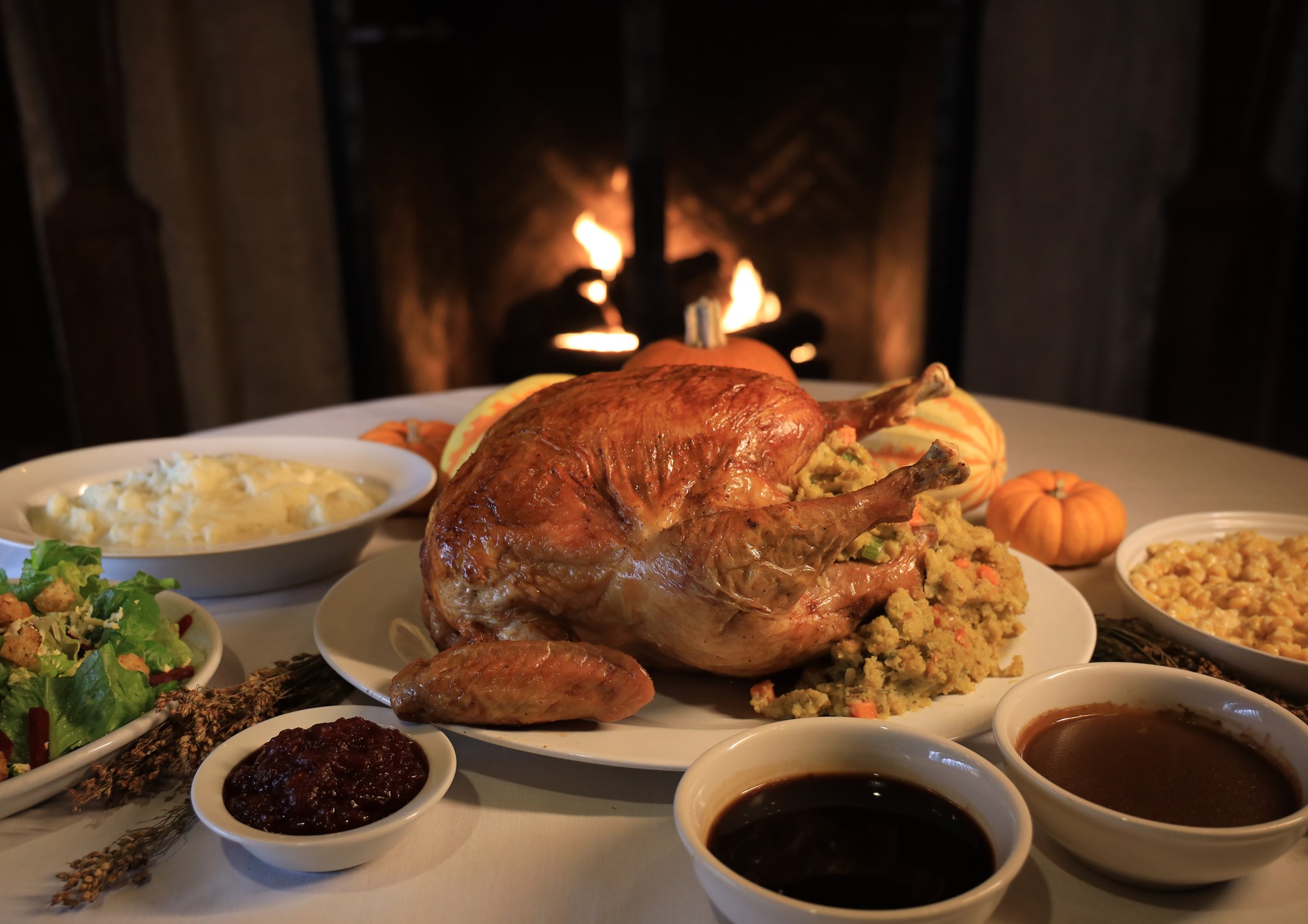 Full turkey with stuffing pouring out of it sitting on a round platter centered on a table with side dishes of salad, mashed potatoes, cranberry sauce, gravy and more surrounding it.