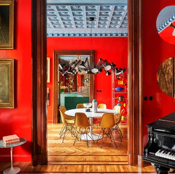 a view from the living room with a baby grand piano at right and a sofa with old paintings hung above it leads into the dining room with a round white dining table and yellow chairs, both rooms with cherry red painted walls