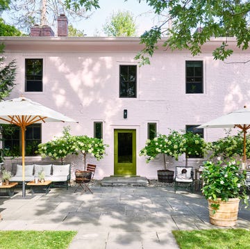 the terrace of a pink two story house has stone flooring, a seating area with sofas and an umbrella at left and a dining table and chairs and umbrella at right, terra cotta urns, and a green door to the house