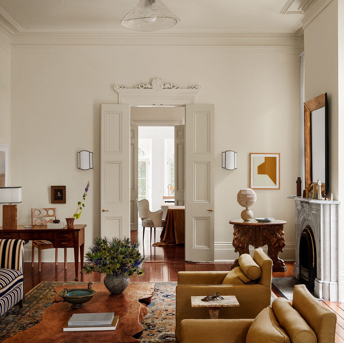 in a living room is a floor lamp and a long striped sofa sits opposite two upholstered chairs, a wood cocktail between, a desk and chair with a table lamp, a round table with three carved legs, sconces flanking the entry