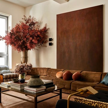 a living room with a long brown suede sofa and a large brown hued artwork above it  cocktail table with books and vases  two 