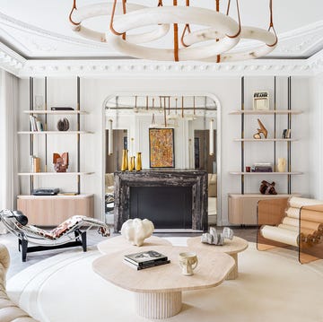 a white room, a trio of circular pendants, semicircle sofa in tufted beige leather, round rug, three cocktail tables, lounge chair, acrylic chair with cylindrical seat, open bookshelves, black marble fireplace with mirror