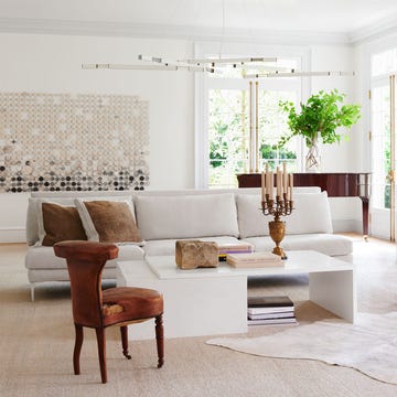 in a white living room is a white sofa, two side chairs, a white cocktail table with books and candelabra, white sideboard, a grand piano, white chandelier with brass arms, a large artwork on back wall, white carpet