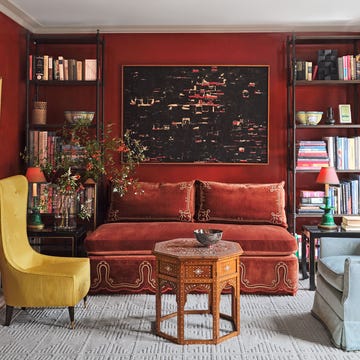 a deep red drawing room has a high back yellow chair, a velvet banquette with decorative cord work flanked by shelves of books and curios, a light blue upholstered chair, and an octagonal cocktail table