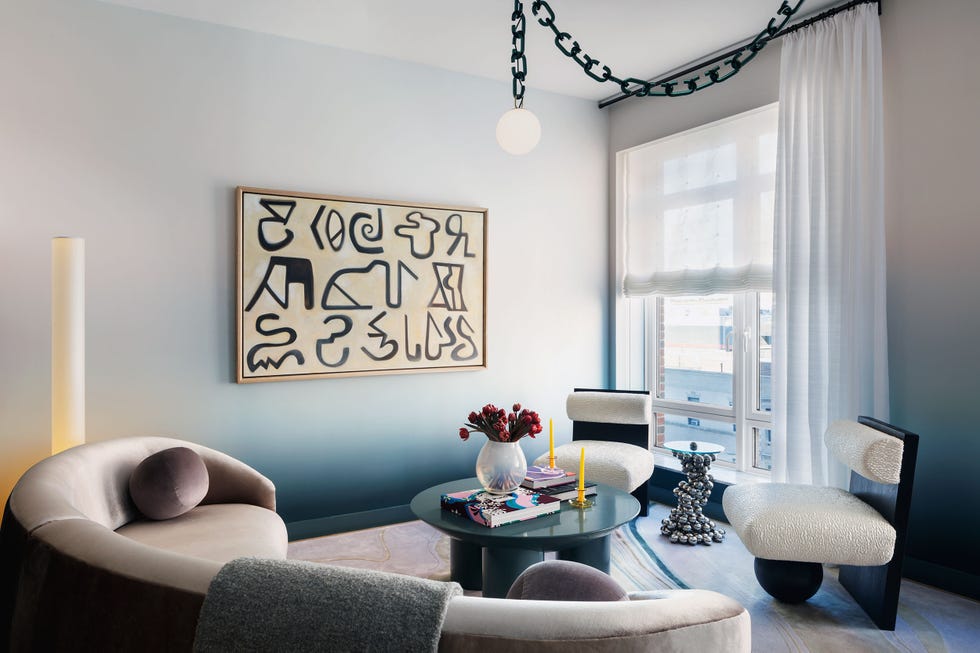 living room with ombre painted walls in shades from palest white blue to darker blue at the base along with a horizontal piece of art with black hieroglyphic type characters on it and a curved sofa in a off gray fabric and two armless chairs with column base and a matte blue round table at center